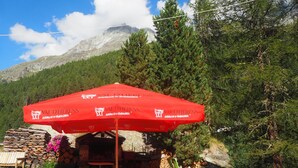 Un paraguas rojo con la marca 'VAL D'HERENS', un edificio de piedra y una montaña al fondo.