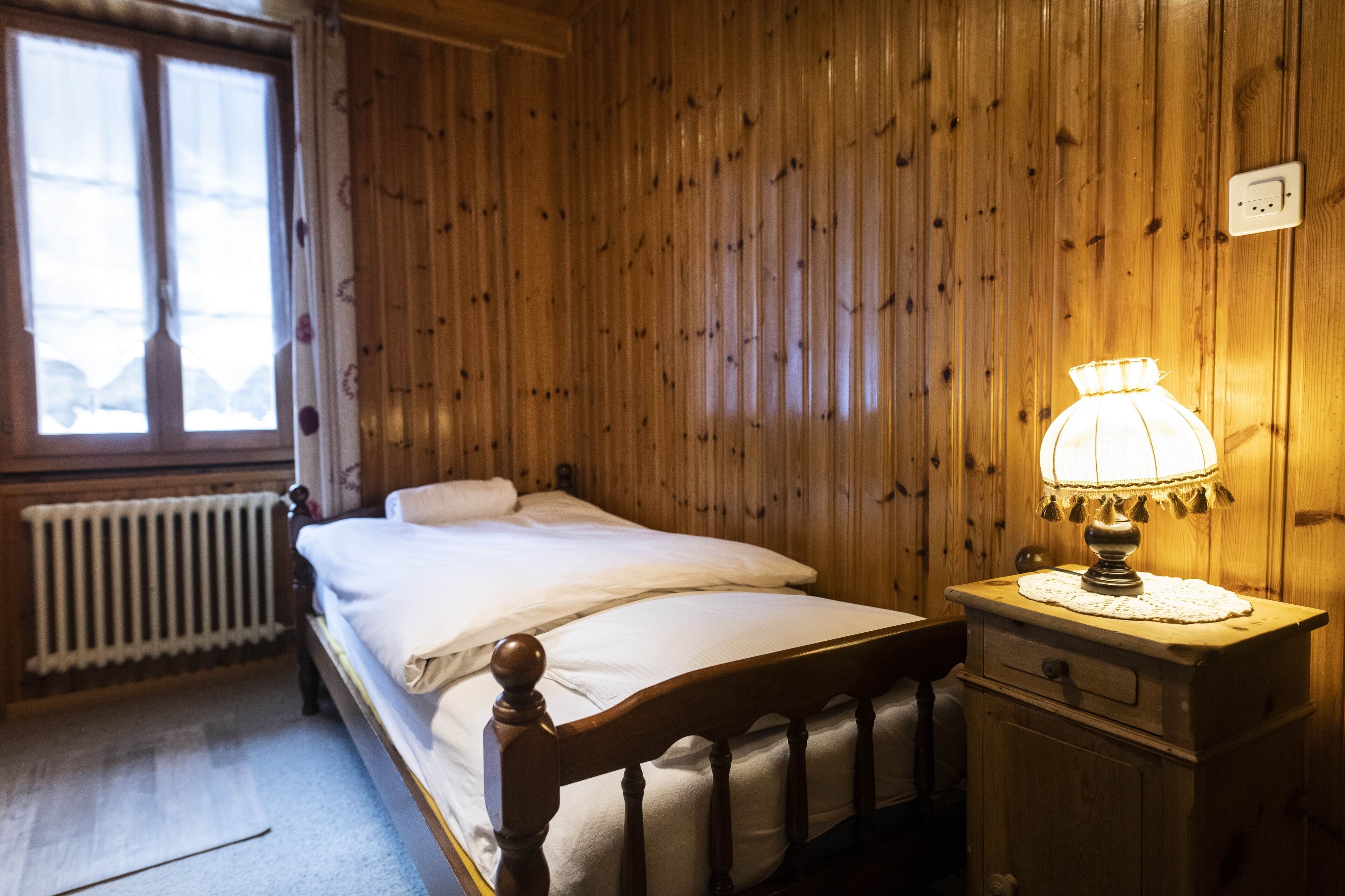 A wooden-paneled room with a bed, a nightstand with a lamp, and a window with curtains.