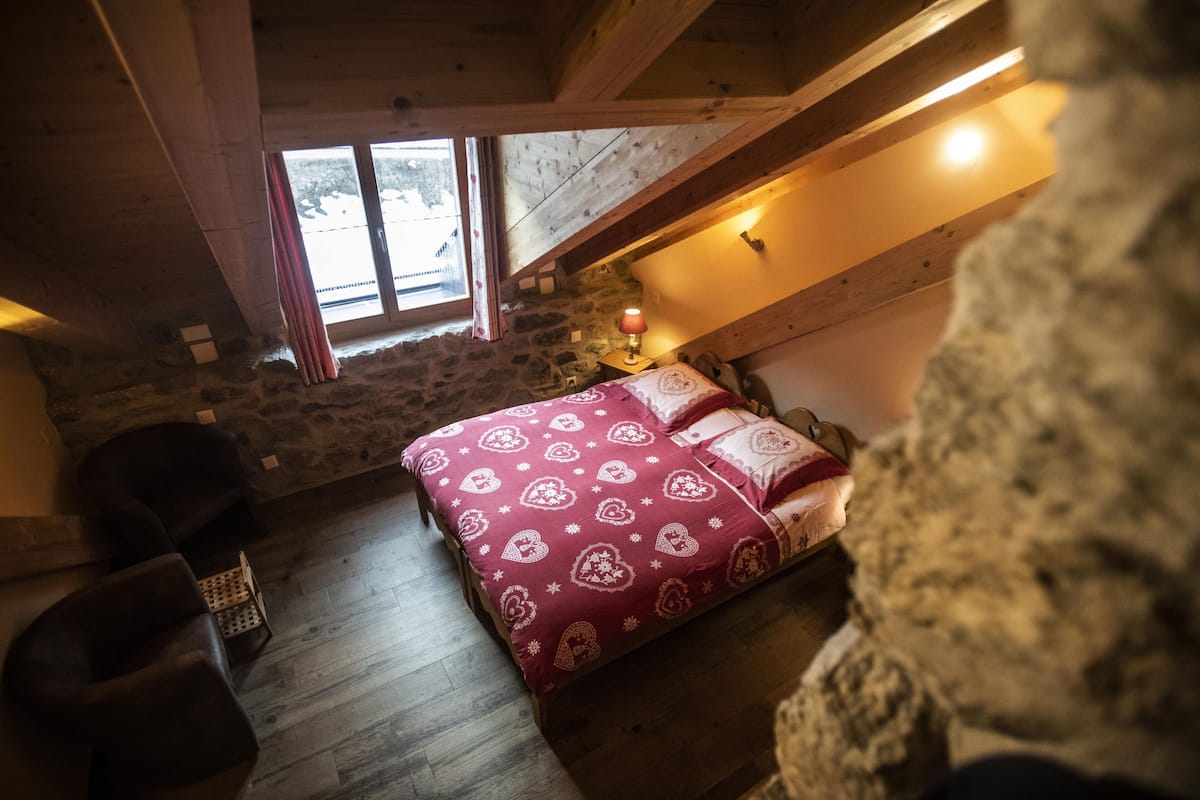 Un dormitorio acogedor en el ático con pared de piedra, techo de madera, cama con colcha estampada, mesita de noche con lámpara y ventana con vistas a árboles.