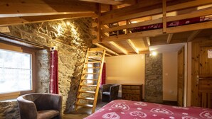 Un dormitorio con techo de madera, paredes de piedra, cama con colcha estampada, una silla, un escritorio y una escalera que lleva a una zona superior.