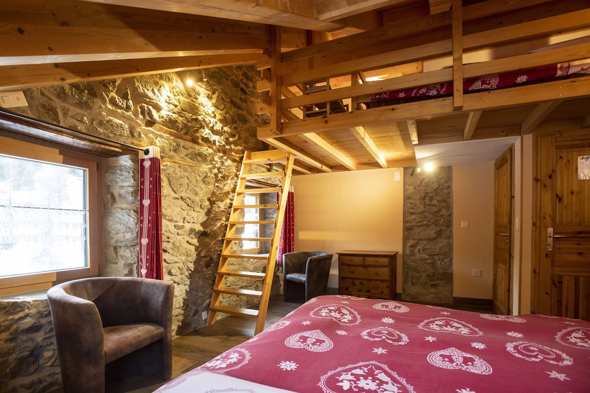Un dormitorio con techo de madera, paredes de piedra, cama con colcha estampada, una silla, un escritorio y una escalera que lleva a una zona superior.