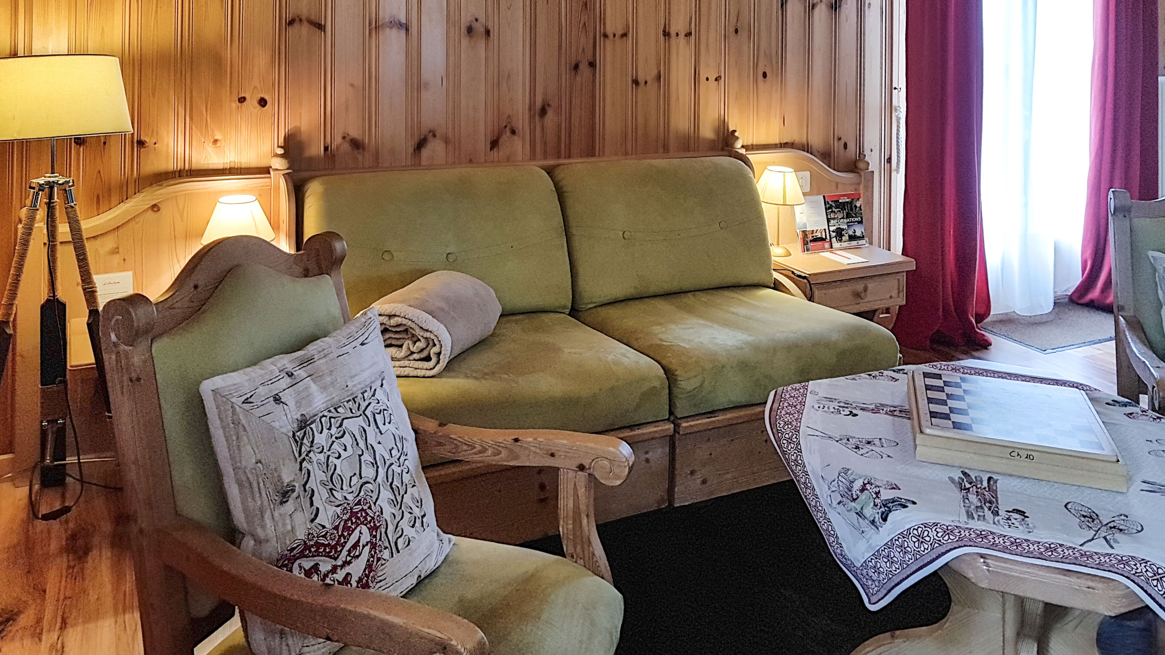 Una habitación acogedora con un sofá verde, un sillón de madera con un cojín estampado, una mesita con un tablero de ajedrez y una lámpara sobre una mesita auxiliar de madera.