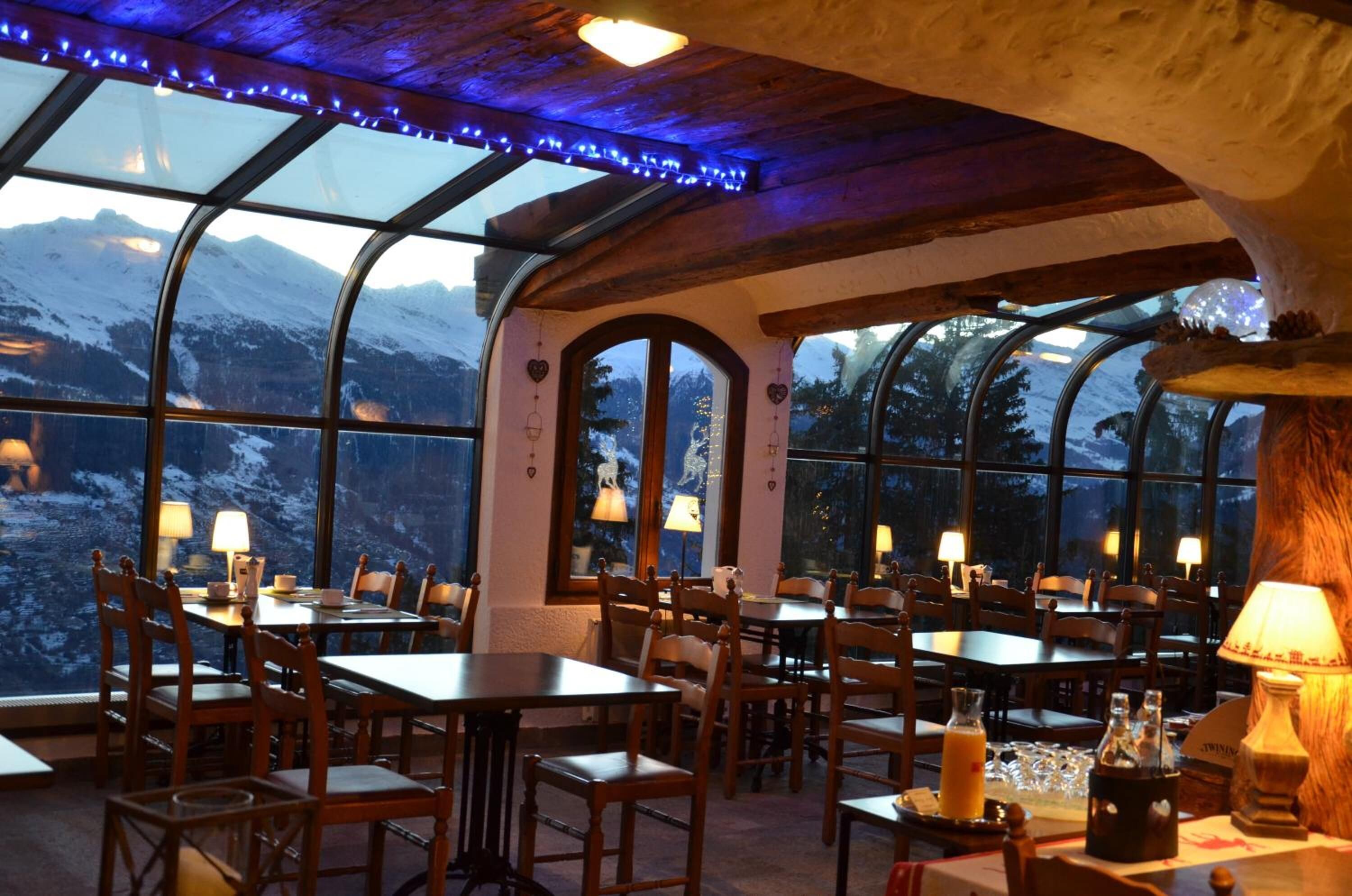 Un restaurante con mesas y sillas dispuestas para comer, amplias ventanas que ofrecen vistas a montañas nevadas y un techo de madera con vigas a la vista.