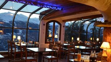 Un restaurante con mesas y sillas dispuestas para comer, amplias ventanas que ofrecen vistas a montañas nevadas y un techo de madera con vigas a la vista.