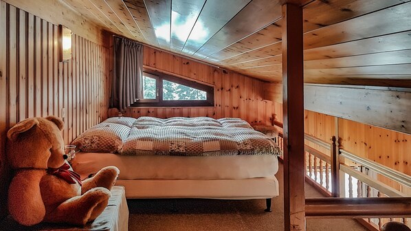 A wooden cabin bedroom with a large bed, a window, a teddy bear, and a staircase.