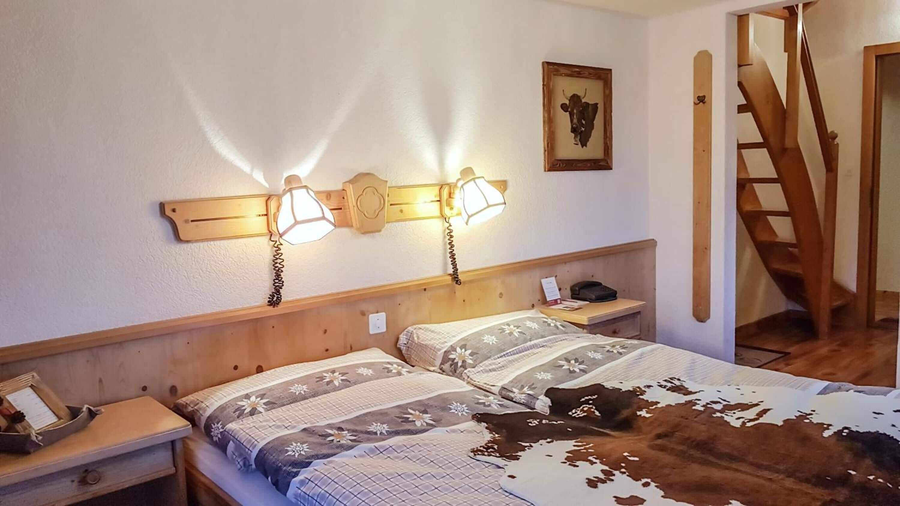 A room with two beds, wooden headboards, bedside tables with lamps, a framed picture, and a wooden staircase.