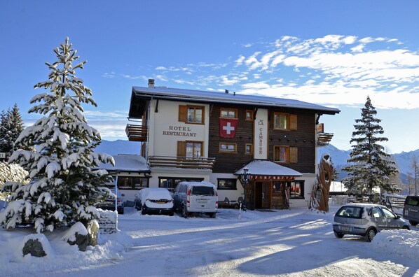 A hotel with a sign reading "HOTEL RESTAURANT" and a red cross symbol. There are cars parked in front, and snow-covered trees around. - Hôtel Restaurant LA CAMBUSE (Vex)