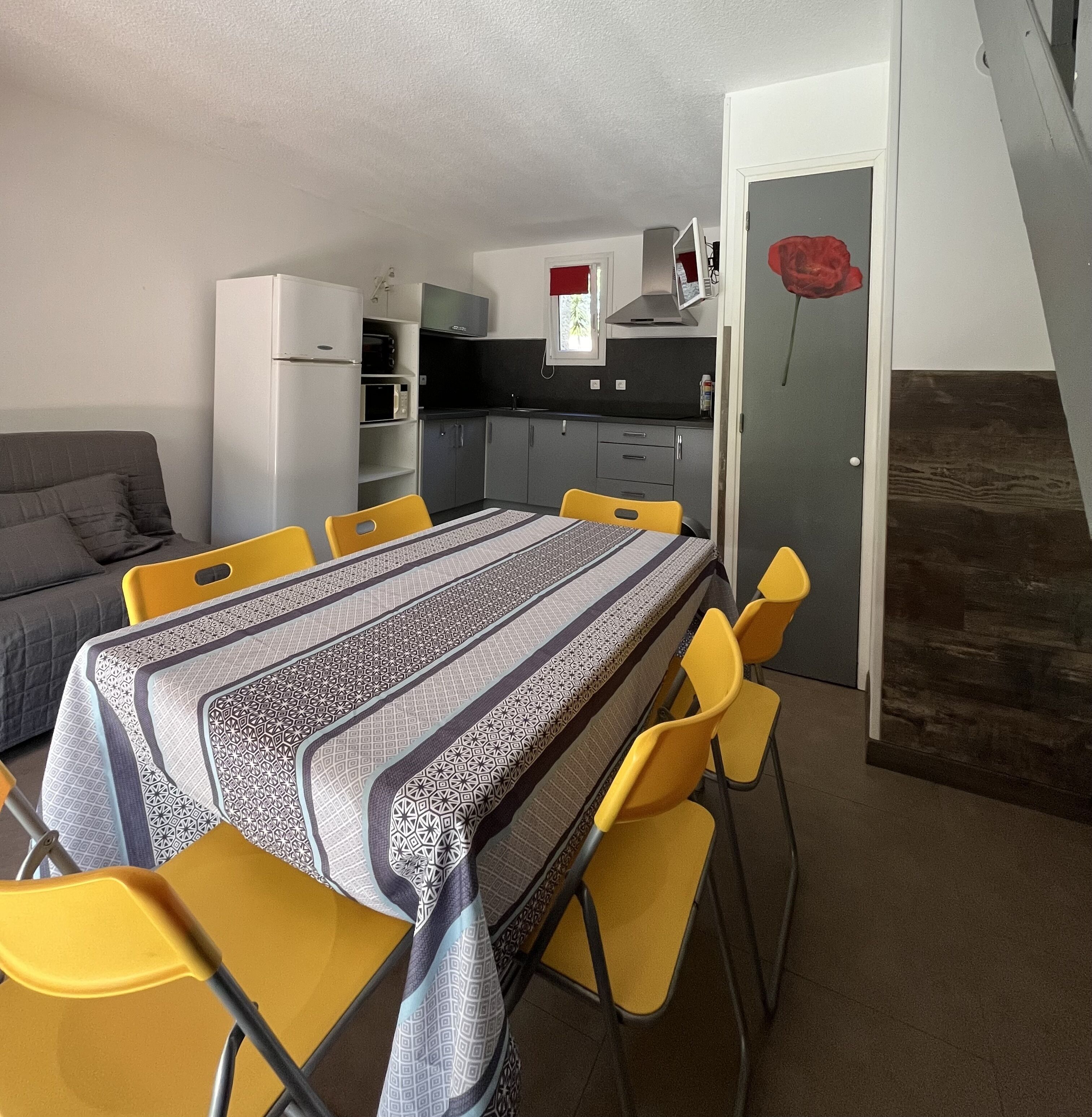 A compact living space with a dining table and chairs, a kitchenette, a gray sofa, and a staircase leading to an upper level.