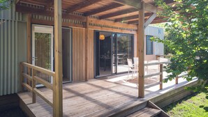 Una terraza de madera con barandilla, puerta de cristal y zona de estar con sillas.