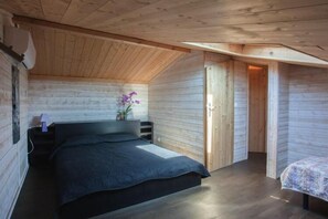 A bedroom with a wooden sloped ceiling, a bed with a dark-colored bedspread, a nightstand with a lamp, and a small potted plant with purple flowers.