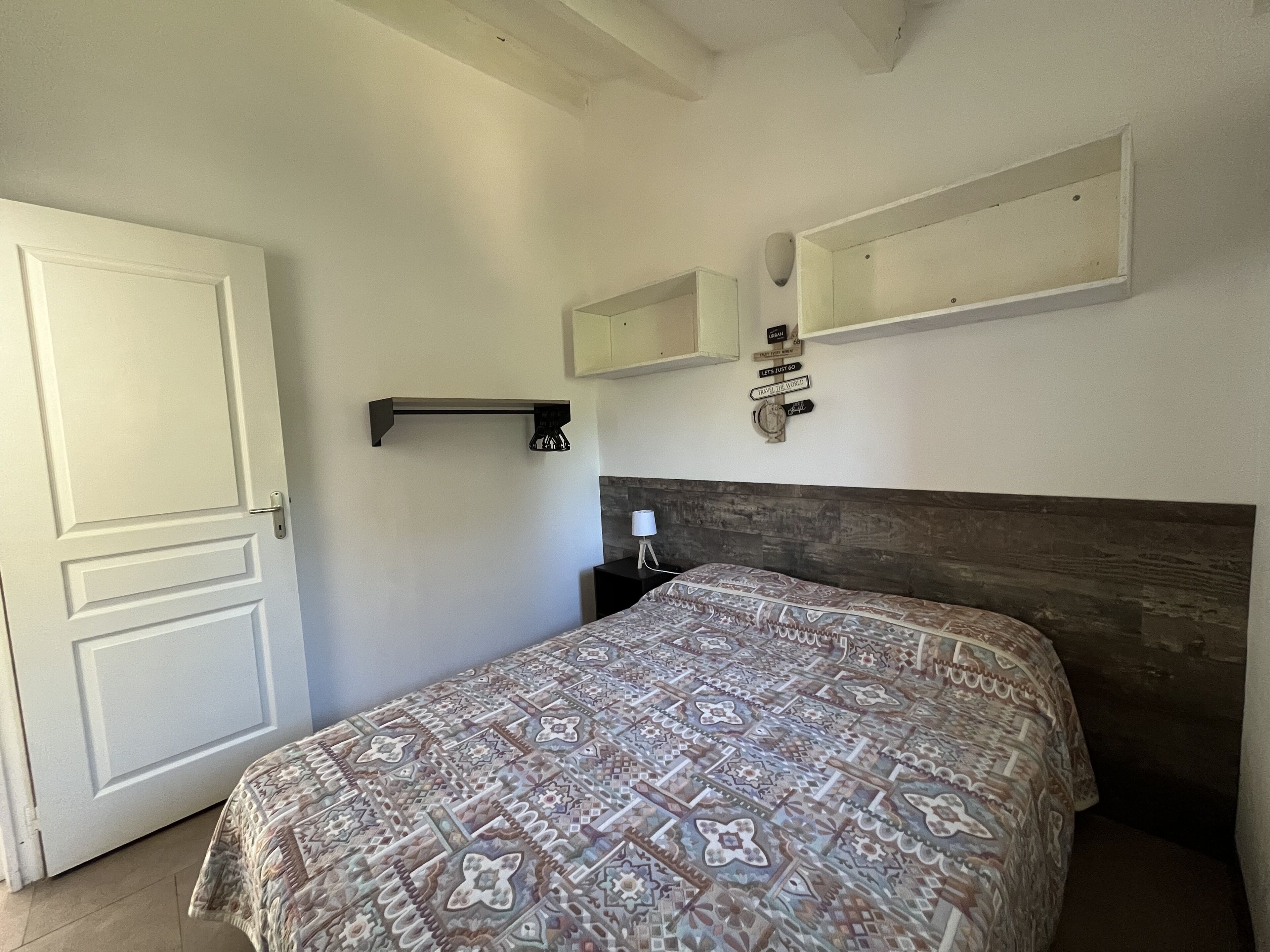 A bedroom with a patterned bedspread, a white door, a bedside table with a lamp, and a wall-mounted shelf with decorative items.