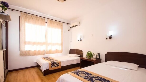 A hotel room with two beds, a window with curtains, a bedside table with a lamp, and an air conditioning unit on the wall.