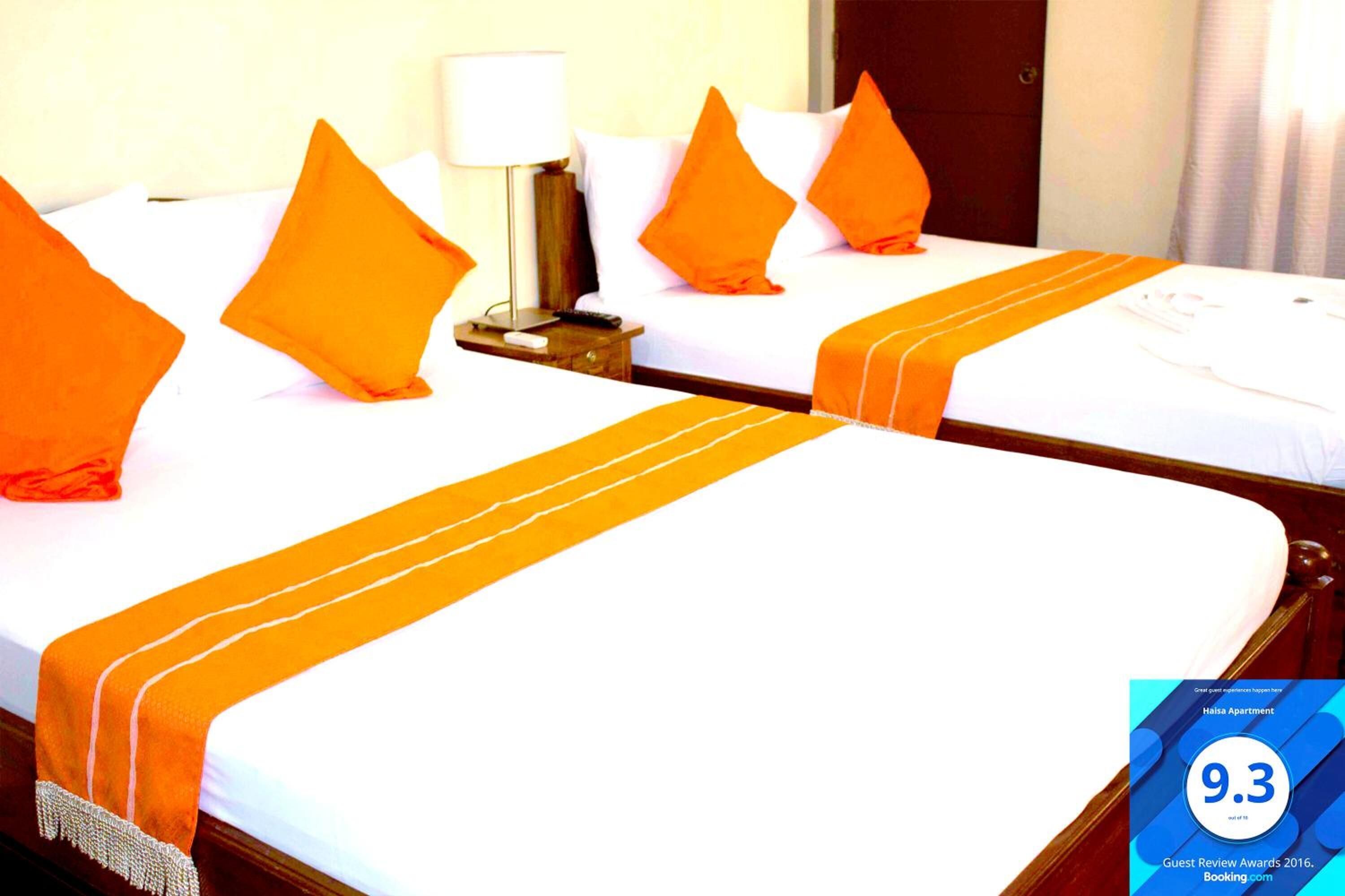 A hotel room with two beds, each with an orange bedspread and pillows. There is a nightstand with a lamp and a remote control.