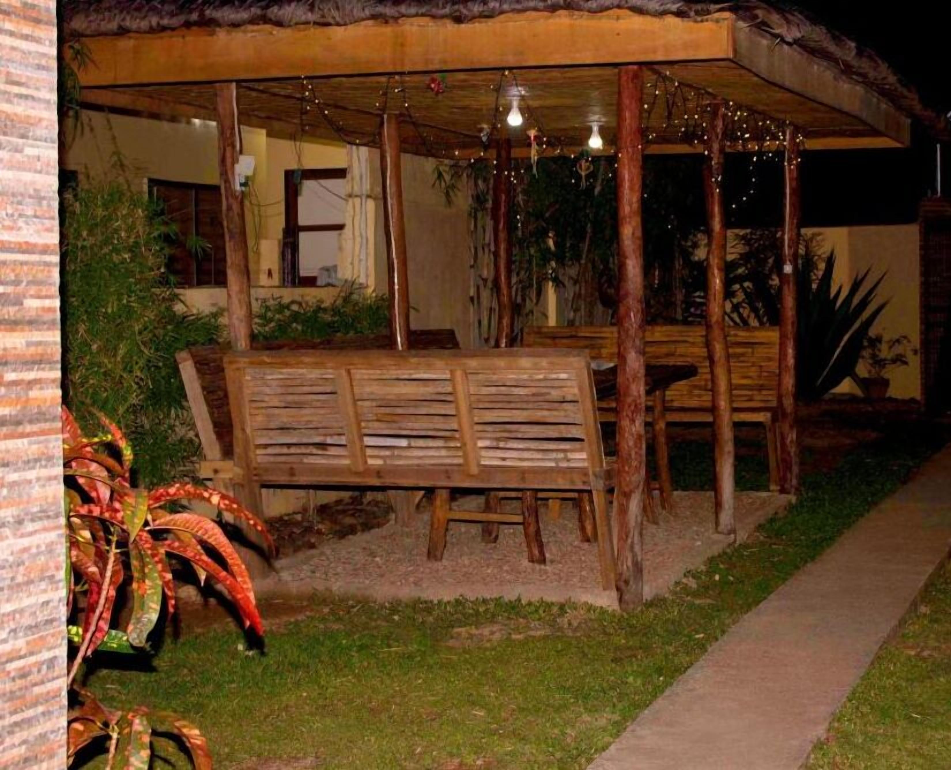 A wooden bench under a thatched roof structure with string lights, surrounded by greenery and a pathway.