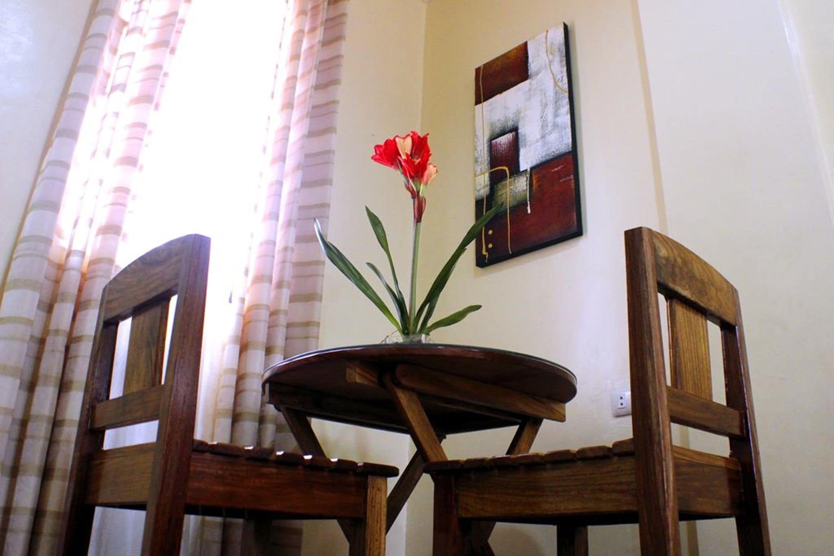 A wooden table with a vase of red flowers, two wooden chairs, and a wall-mounted abstract painting.