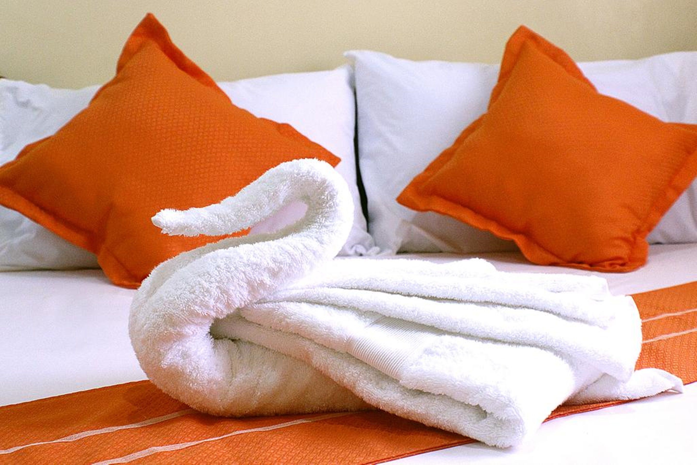 Ein ordentlich gemachtes Bett mit weißen und orangefarbenen Kissen und einem weißen Handtuch, das zu einer Schwanform geformt ist.
