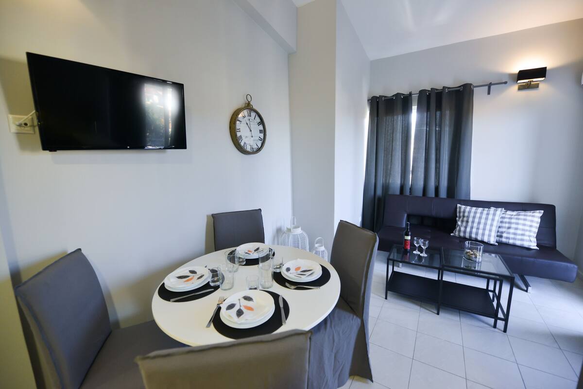 A modern living room with a flat-screen TV, a clock, a dining table set for four, and a sofa with a checkered pillow.
