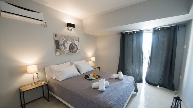 A neatly made bed with a gray bedspread, white pillows, and two towels. There is a small table with a lamp and a tray of fruit. The room has a large window with curtains, a wall-mounted air conditioner, and a piece of abstract art. - Regalo Apartments (Lefkada)