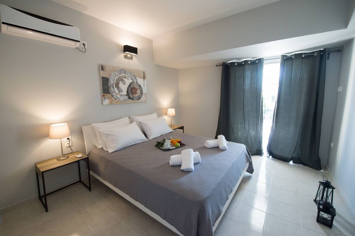A neatly made bed with a gray bedspread, white pillows, and two towels. There is a small table with a lamp and a tray of fruit. The room has a large window with curtains, a wall-mounted air conditioner, and a piece of abstract art.