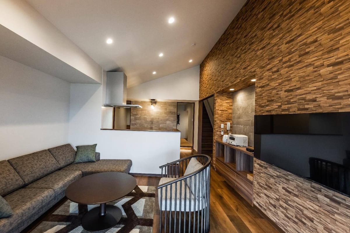 A modern living room with a sectional sofa, a round coffee table, a staircase, and a kitchen area with a built-in oven.