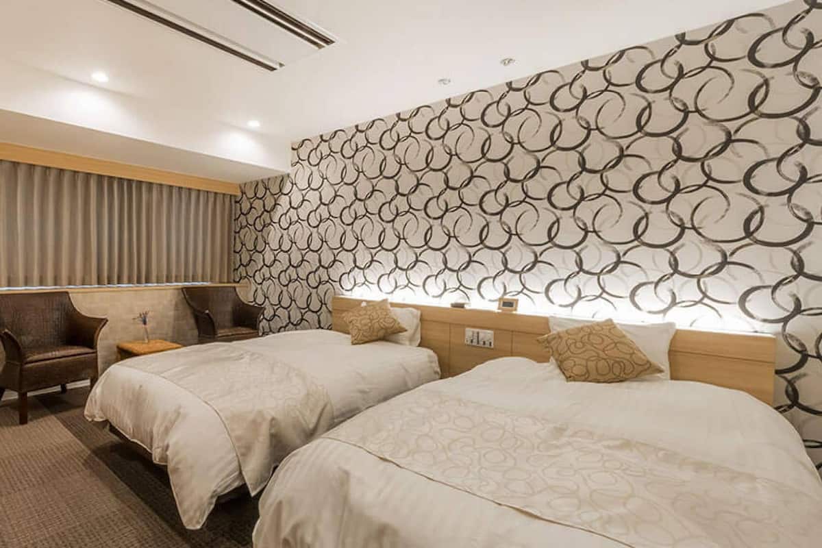 A hotel room with two beds, a desk, a chair, and a patterned wall.