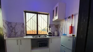 Une cuisine avec des meubles blancs, une cuisinière à gaz, un four à micro-ondes, un réfrigérateur et une fenêtre avec des barreaux.