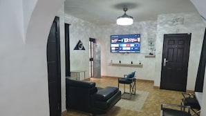 Un salon avec un canapé noir, une table en verre, un téléviseur fixé au mur et une porte.
