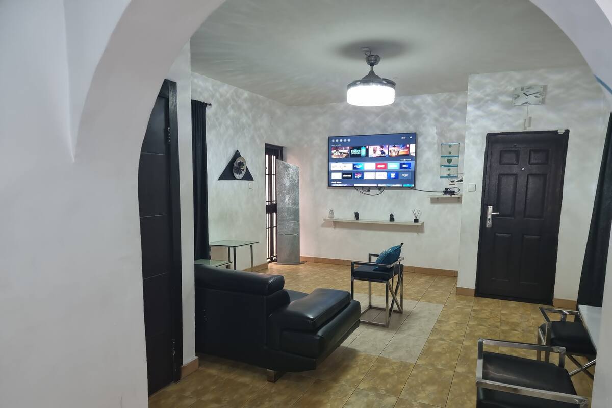 A living room with a black sofa, a glass table, a TV mounted on the wall, and a door.
