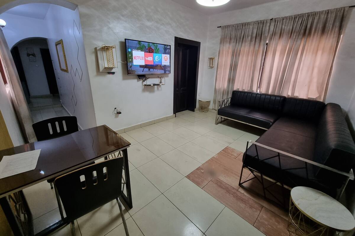A living room with a TV mounted on the wall, a sofa, a dining table with chairs, and a small round table.