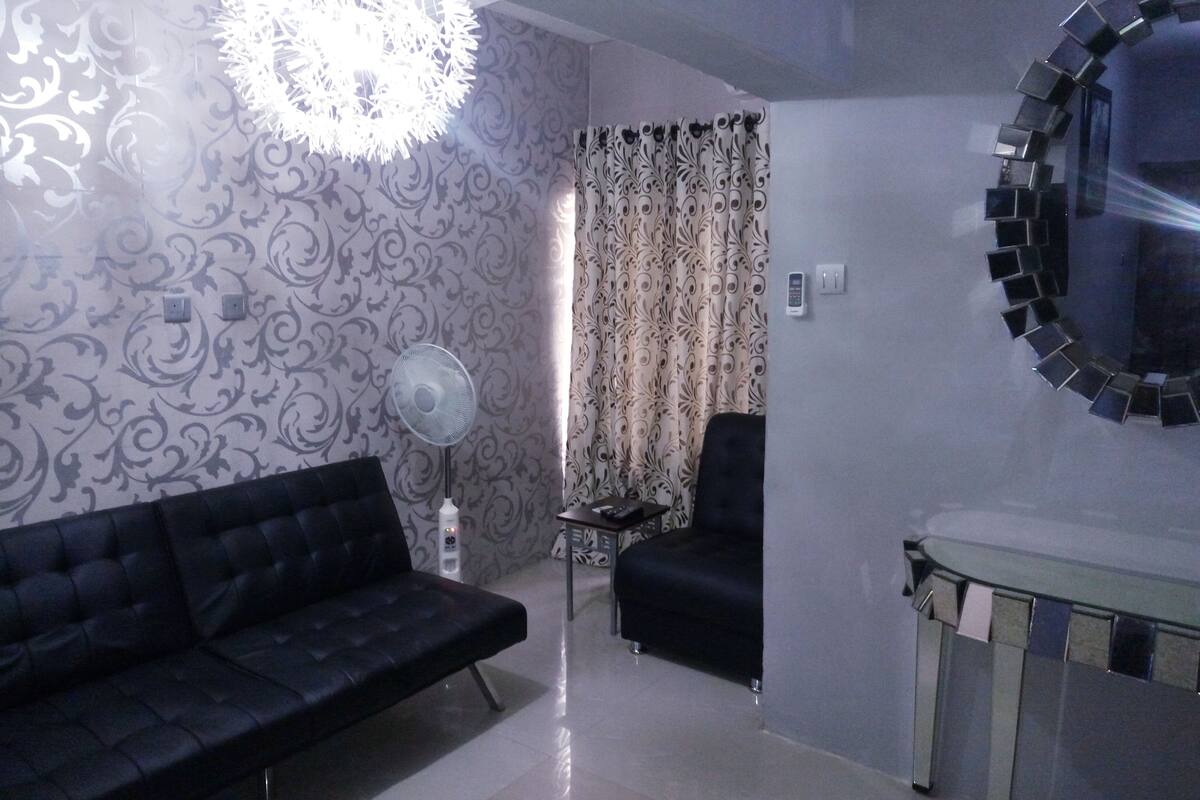A living room with a black sofa, a standing fan, a patterned wall, and a mirror on the wall.