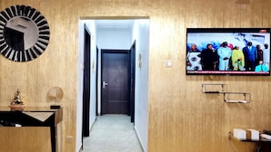 Un couloir avec un mur en bois, un téléviseur diffusant une image d’un groupe de personnes et une porte au fond.