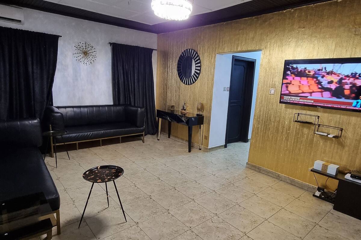 A living room with a black leather sofa, a small round table, a TV mounted on a wooden wall, and a door leading to another room.