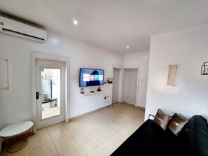 A living room with a flat-screen TV mounted on the wall, a sofa, a small table, and a door leading to another room. - Epic Homes (Lagos)