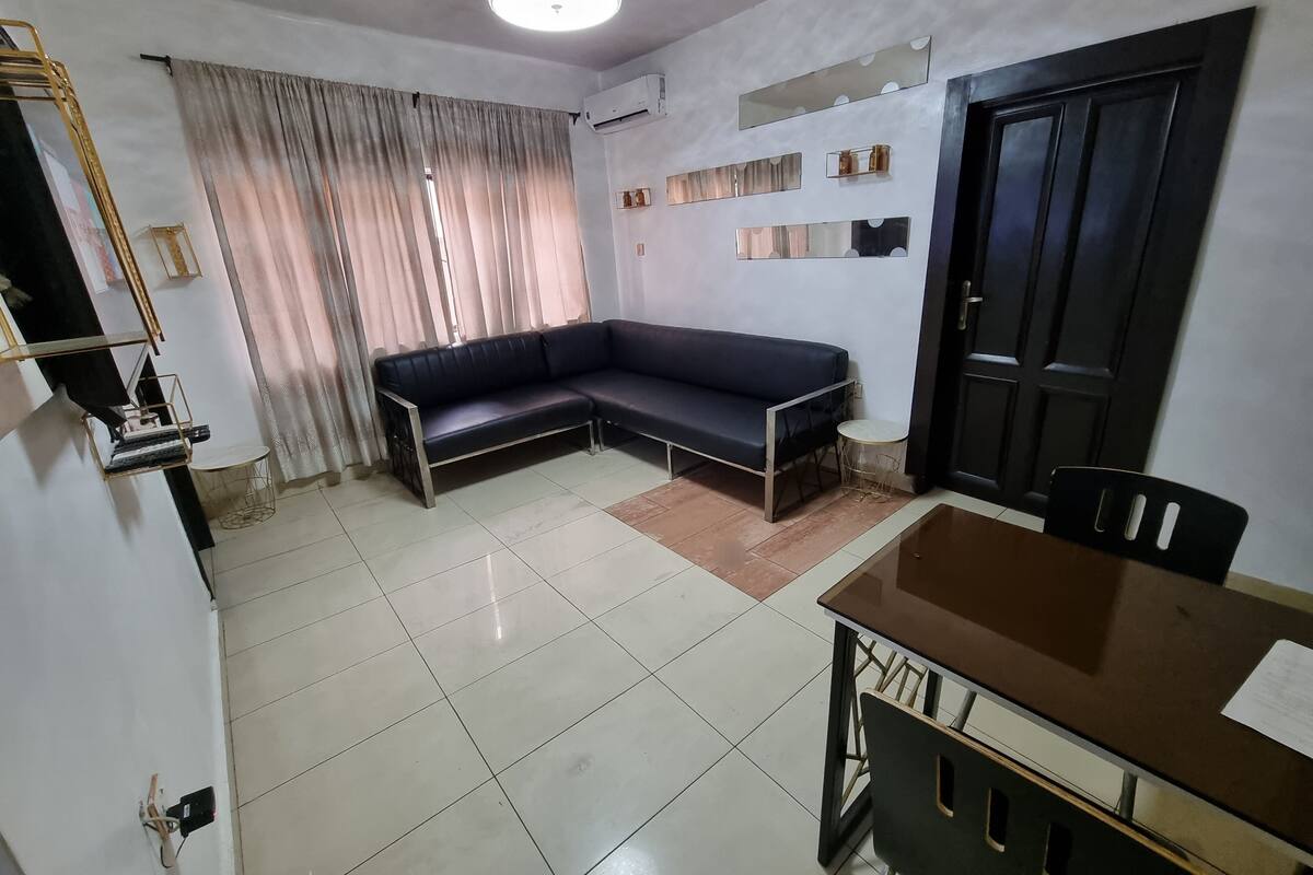 A living room with a sectional sofa, a dining table with chairs, a ceiling light fixture, and a door.