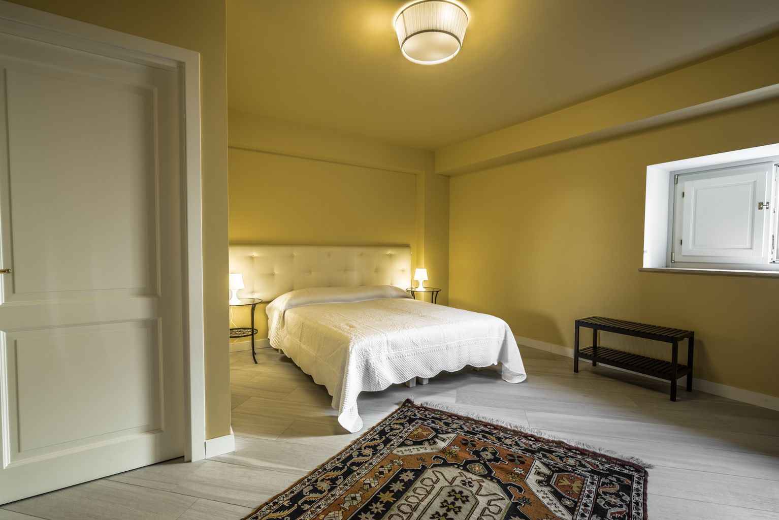 A bedroom with a bed, bedside lamps, a bench, a patterned rug, and a ceiling light.