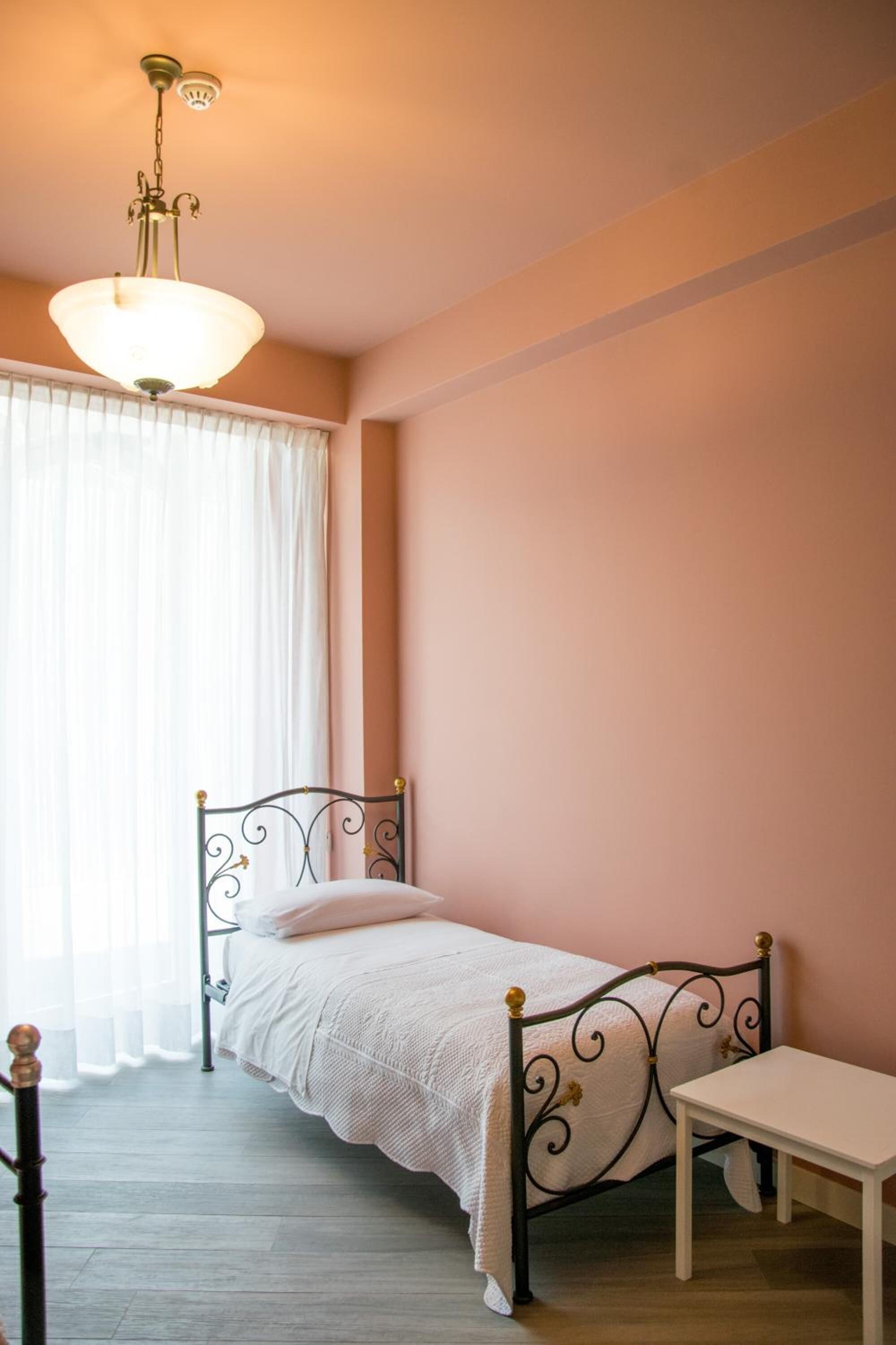 Een enkel bed met metalen frame en een witte matras, een nachtkastje en een hangende lamp in een kamer met roze muren en een houten vloer.