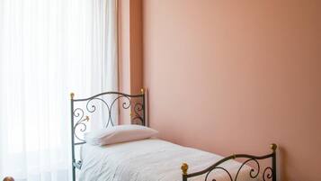 Een enkel bed met metalen frame en een witte matras, een nachtkastje en een hangende lamp in een kamer met roze muren en een houten vloer.