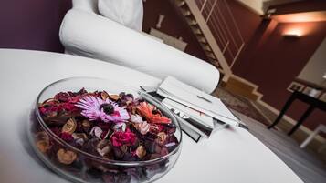 Un tazón de vidrio transparente lleno de flores secas y nueces sobre una mesa blanca, con una almohada blanca y una pila de revistas al fondo.