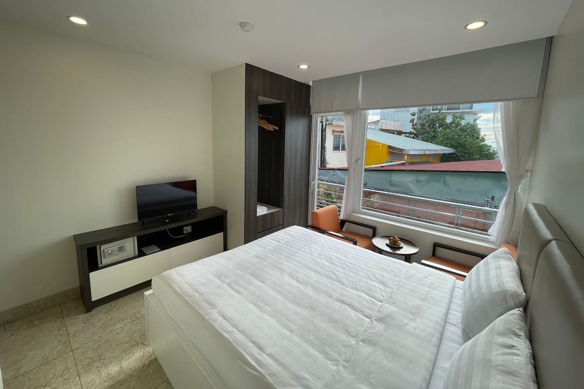 Ein modernes Hotelzimmer mit einem großen Bett, einem Fernseher auf einem Schrank, einem kleinen Tisch mit einer Fruchtschale und einem Fenster mit Blick auf Gebäude.
