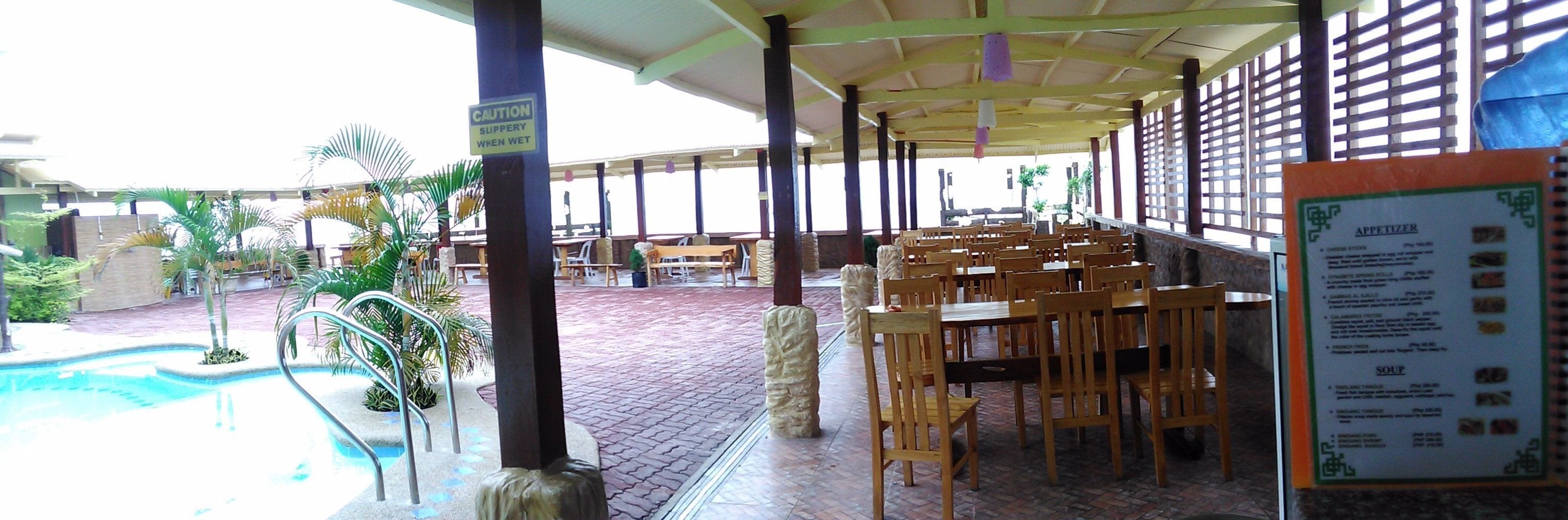 Uma área de estar coberta ao ar livre com mesas, cadeiras, piscina e um quadro de cardápio.