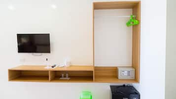 Una habitación con televisión montada en la pared, un estante de madera con una caja fuerte pequeña, un cubo de basura verde y un bolso negro en el suelo.