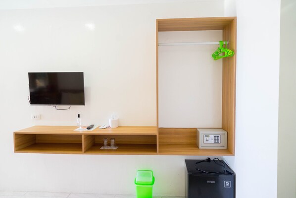 Una habitación con televisión montada en la pared, un estante de madera con una caja fuerte pequeña, un cubo de basura verde y un bolso negro en el suelo.