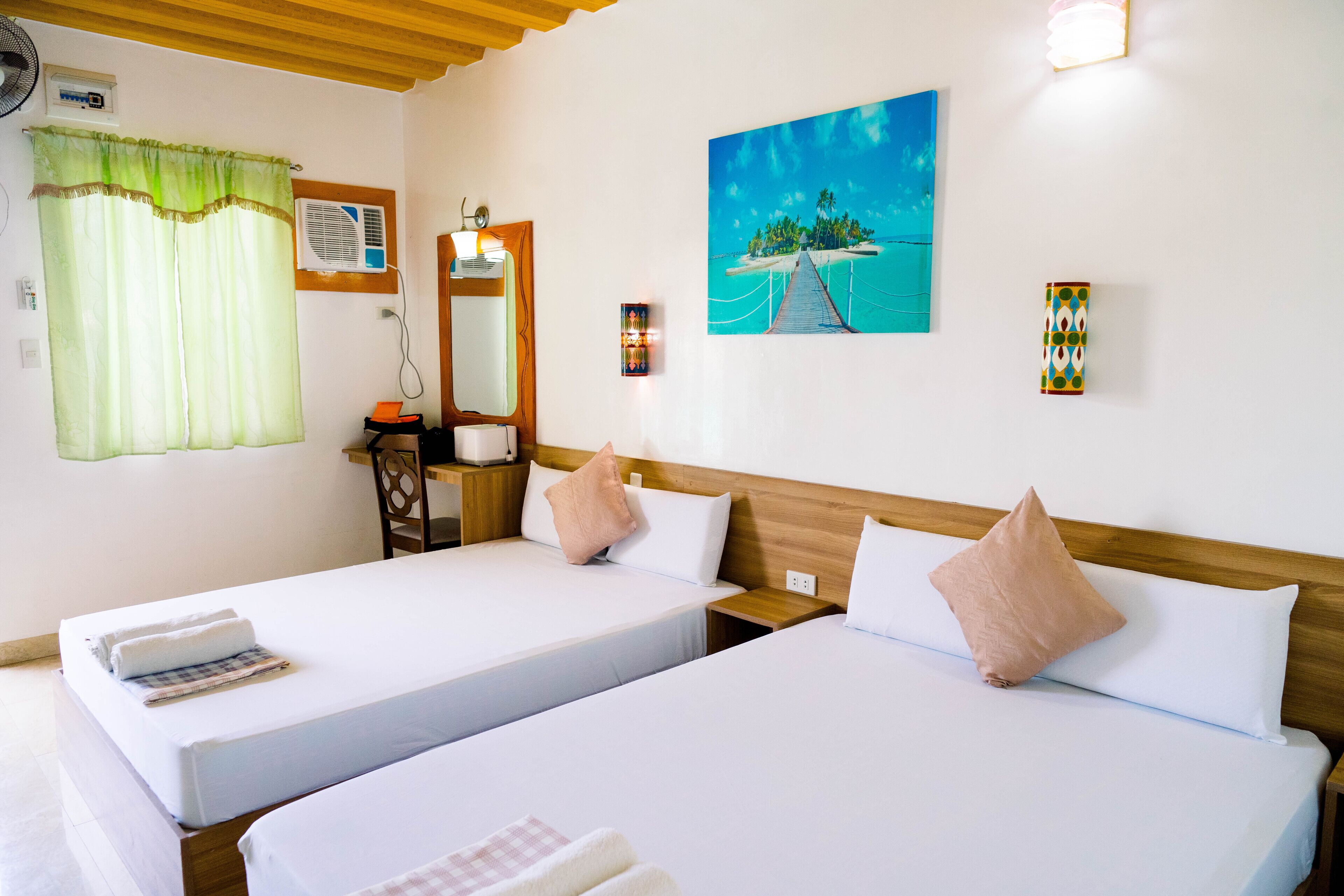 A hotel room with two beds, a desk, a chair, an air conditioner, a painting, and a wall-mounted light fixture.