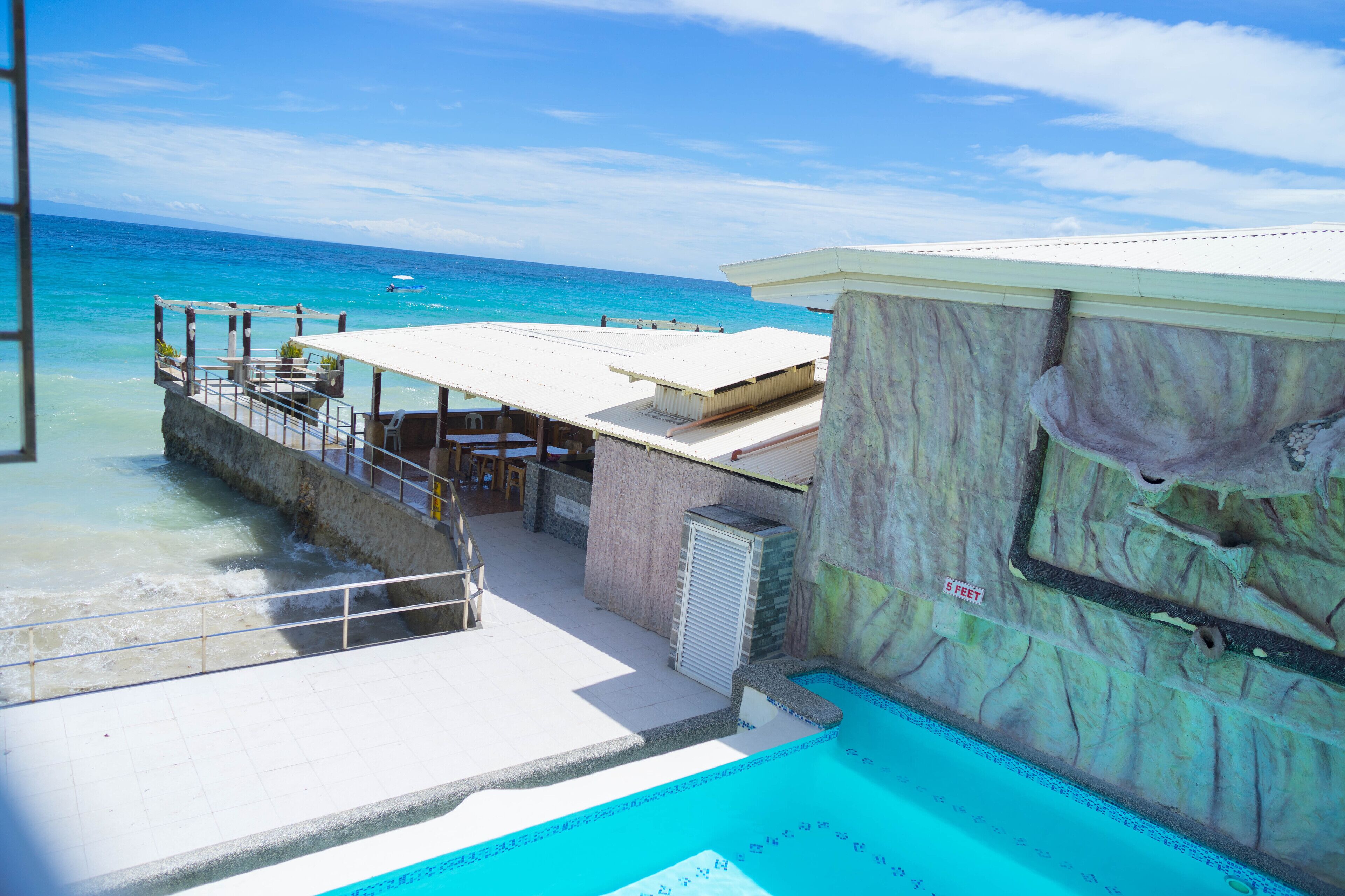 Un área de piscina con vista al océano, una zona de estar cubierta y un edificio con fachada de piedra.