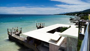 A coastal hotel with a white building overlooking turquoise waters, a pier with seating, and a clear sky.