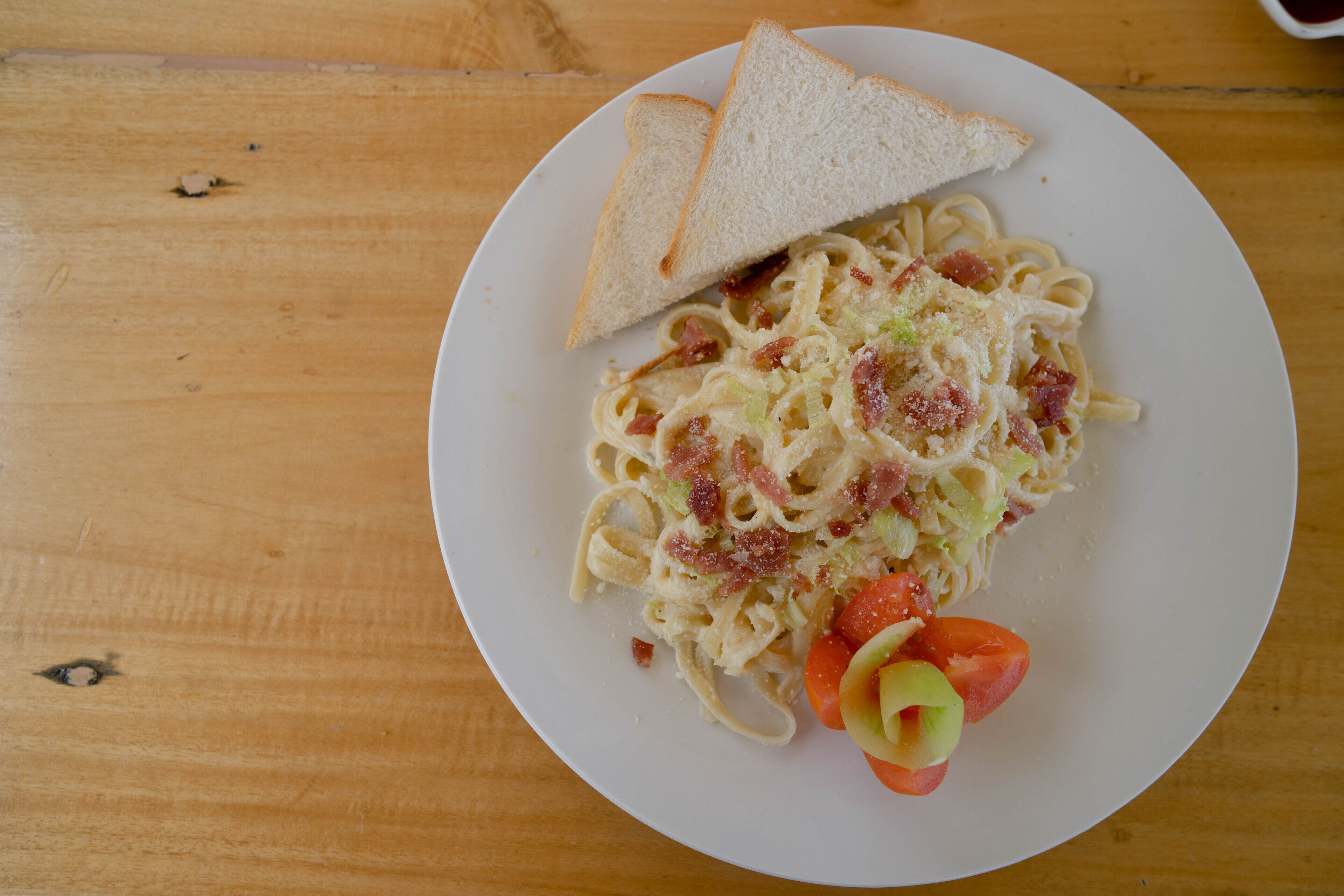 Een bord pasta met spek en plakjes tomaat op een houten tafel.