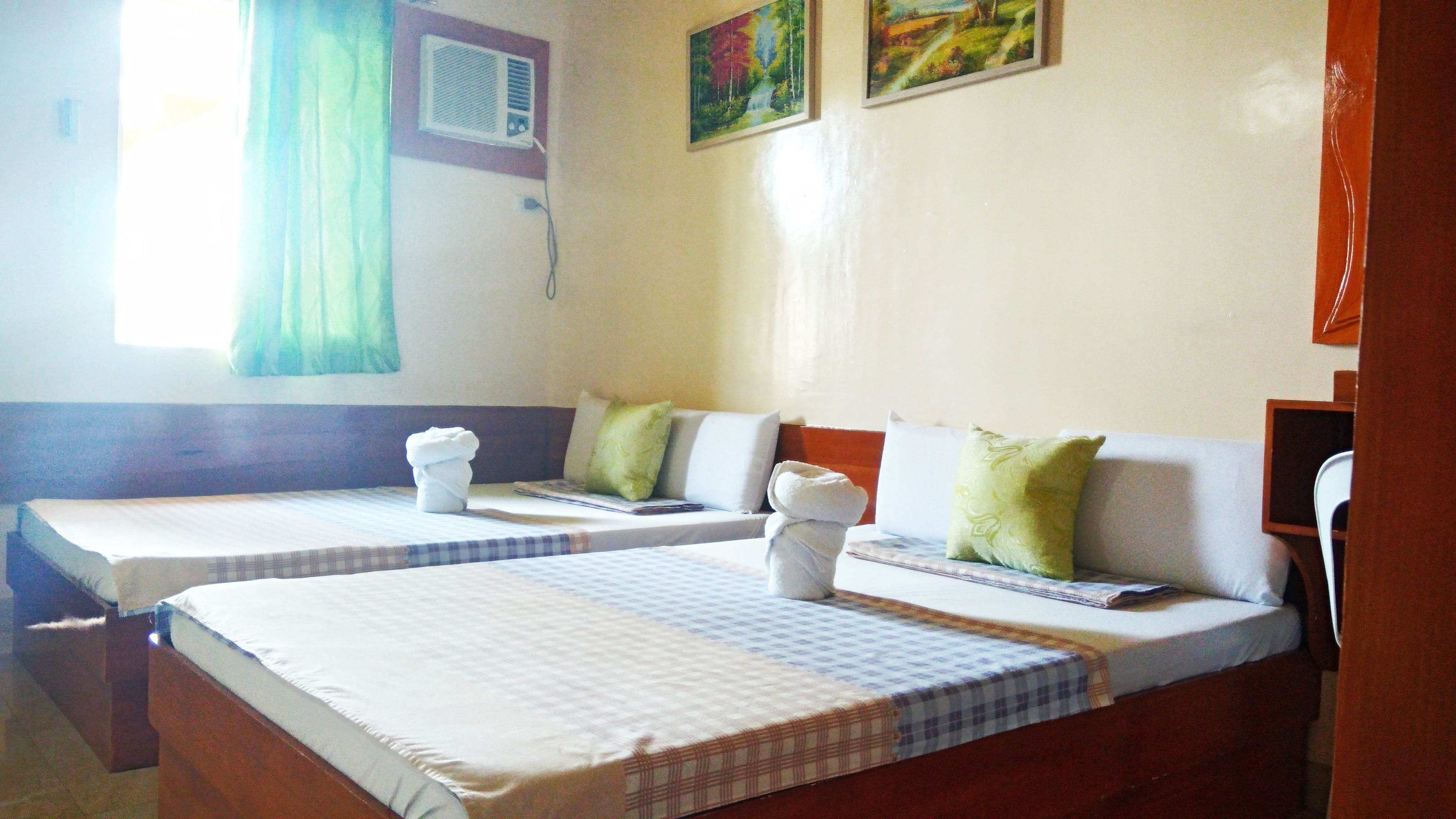A hotel room with two single beds, each with a checkered bedspread and pillows. There is a window with a curtain, a wall-mounted air conditioning unit, and two paintings on the wall.