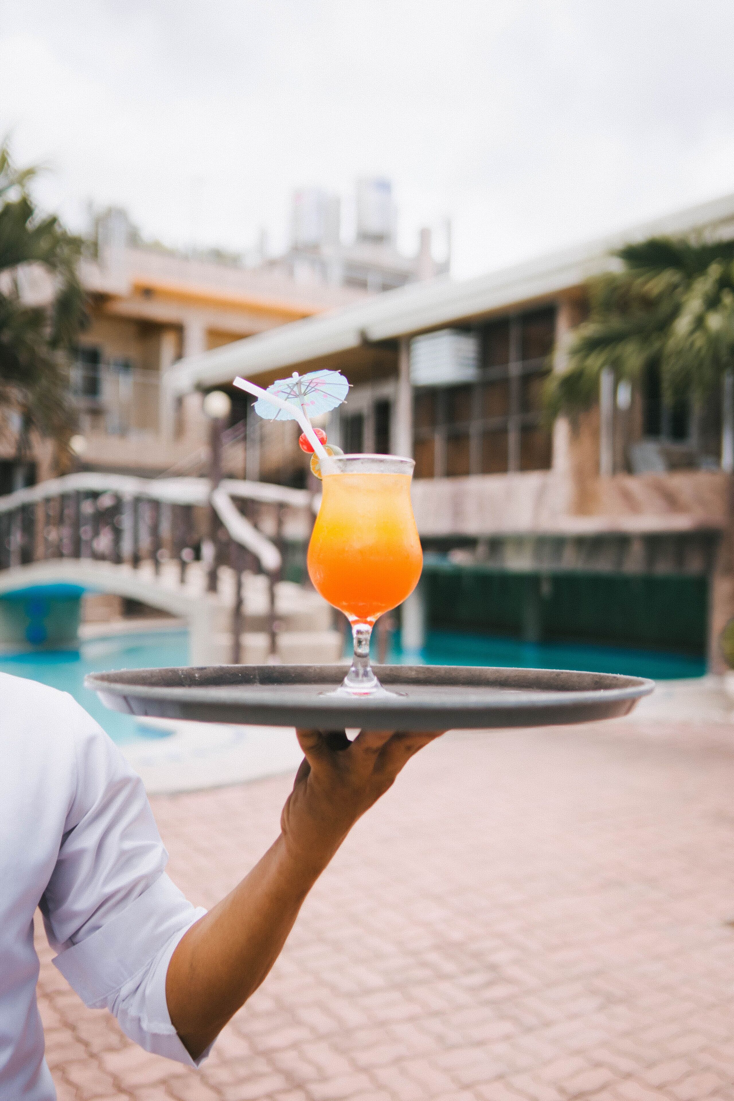 Una persona sostiene una bandeja con un vaso de bebida de naranja y una pajita, cerca de una piscina con un puente al fondo.