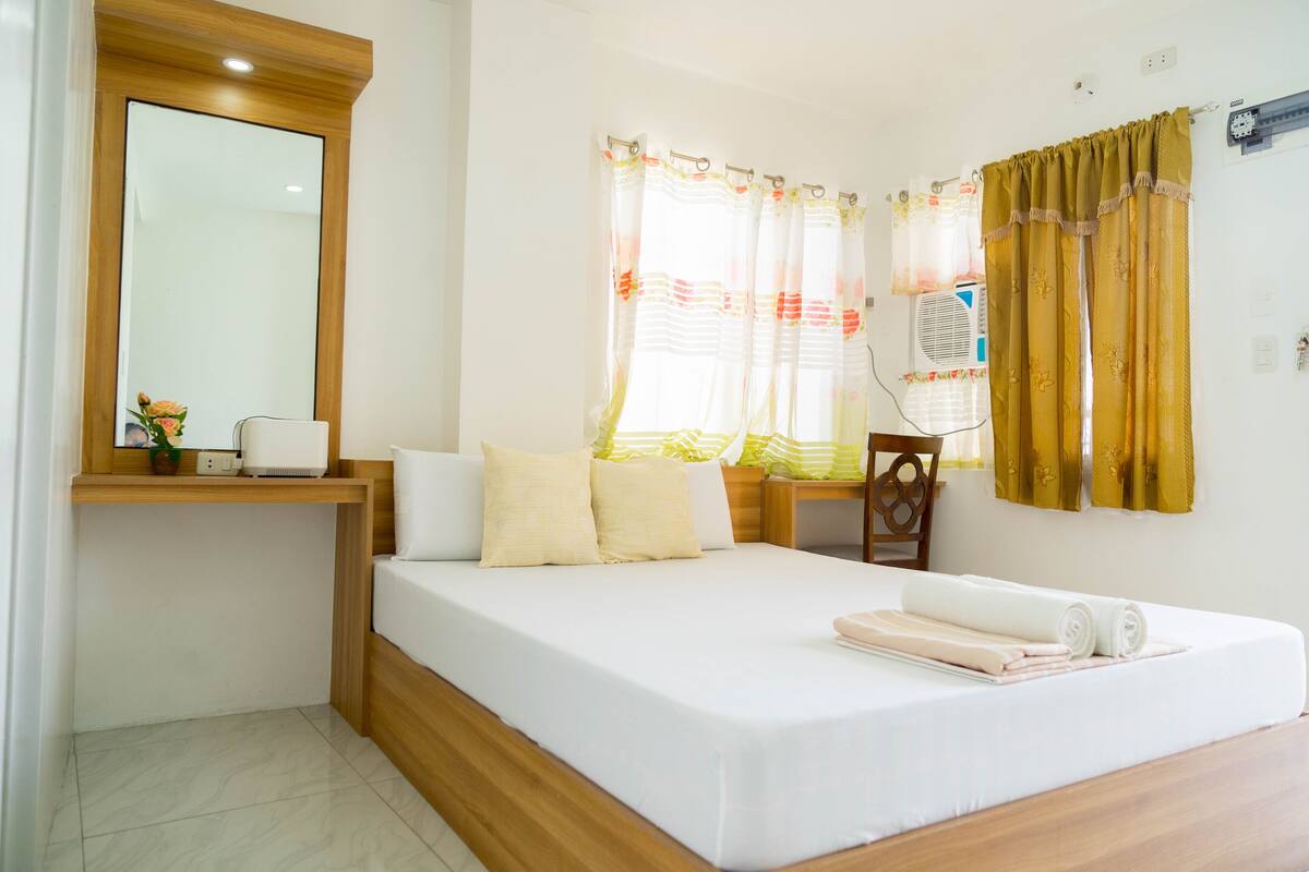 A hotel room with a wooden bed frame, white bedding, a bedside table with a lamp, and a large mirror.