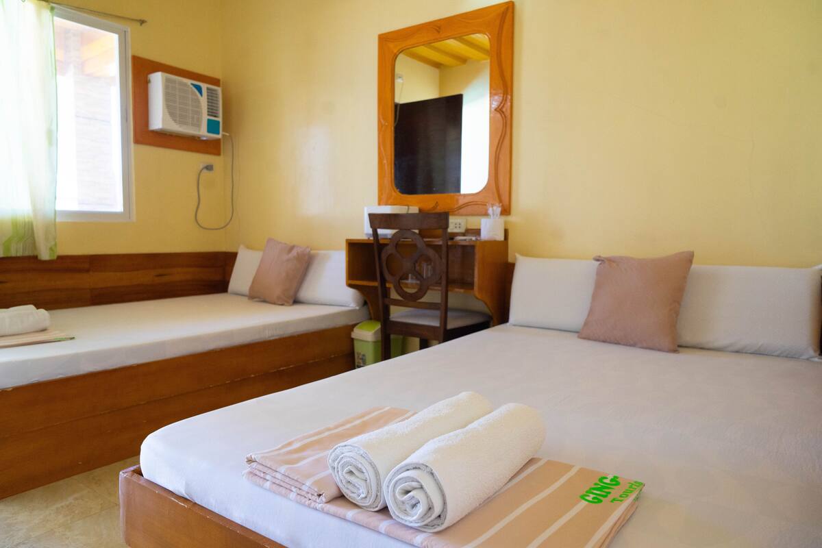 A hotel room with a bed, a desk, and a chair. There is a window with curtains, an air conditioning unit, and a mirror on the wall.
